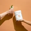Two hands exchange a glass of beige shake with a mint garnish against a peach wall, while a white tub labeled 'Life Shake' is held nearby (Shaklee).