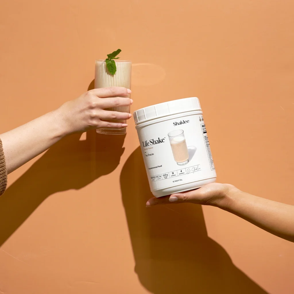 French Vanilla Life Shake Soy Protein Powder Shaklee - Shop Shaklee Australia Two hands exchange a glass of beige shake with a mint garnish against a peach wall, while a white tub labeled 'Life Shake' is held nearby (Shaklee).