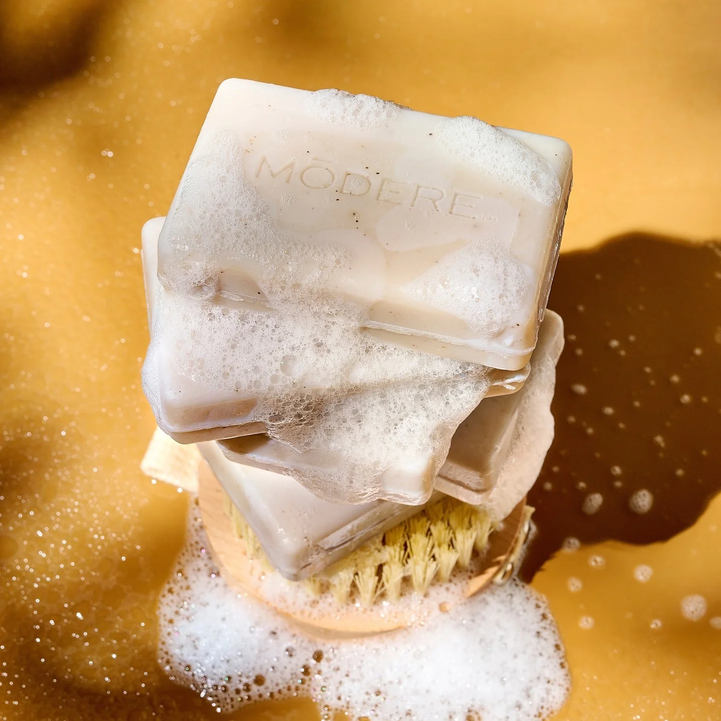 Modere Body Bar Australia - Image 2 - Shop Shaklee Australia Stack of pale soap bars with bubbles, the word MODERE etched on the top bar, resting on a brush in foamy yellow water