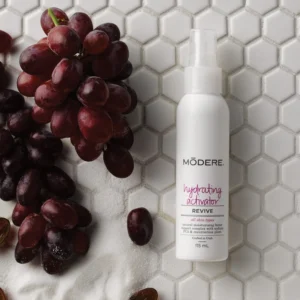 Bunch of red grapes next to a white Modere bottle on a white hex-tile surface, likely a skincare product.
