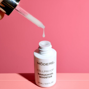 White Modere Cellproof Bakuchiol Booster bottle with a dropper releasing a drop of serum against a pink background.