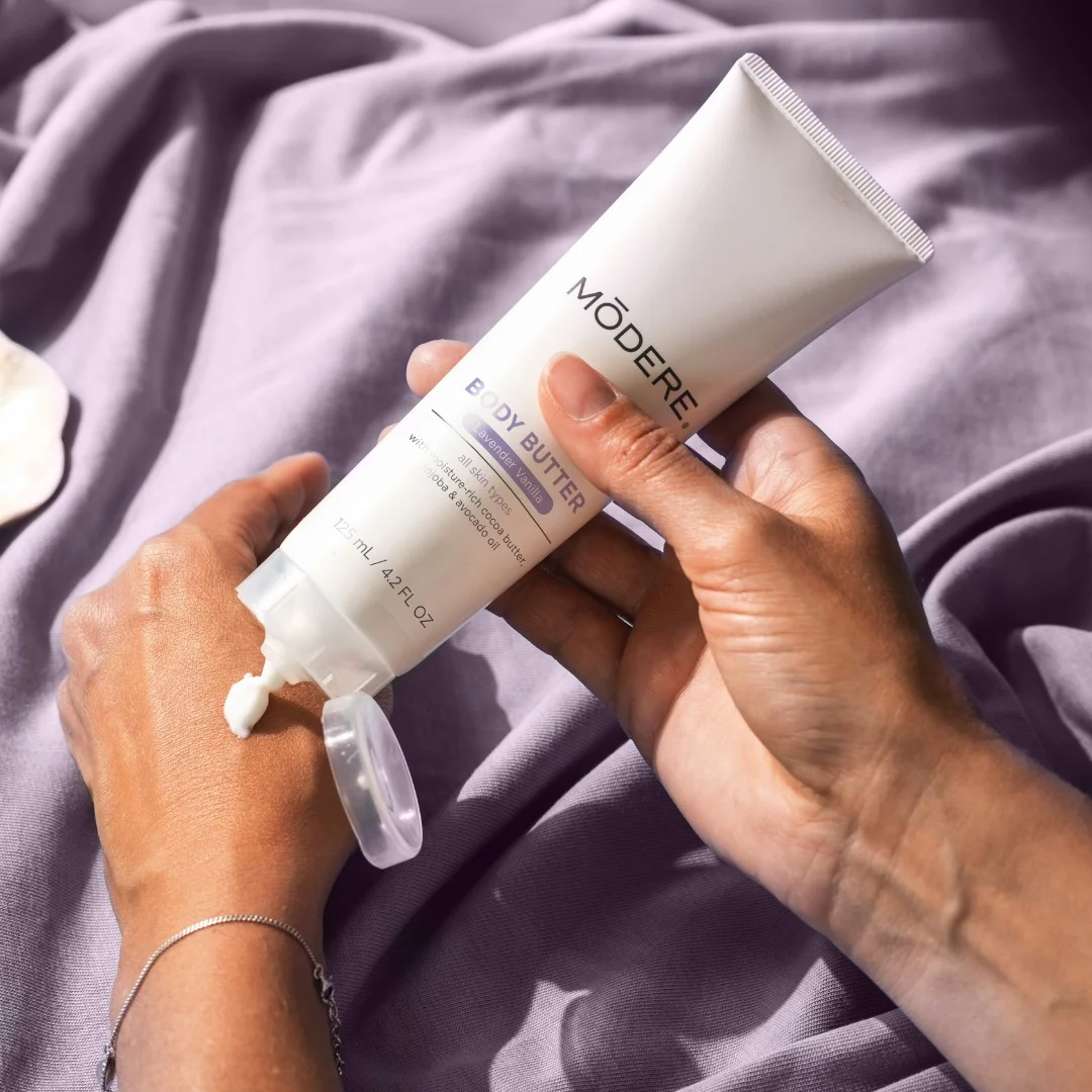 Modere Body Butter Australia - Image 3 - Shop Shaklee Australia Close-up of hands squeezing a white tube labeled 'Body Butter' onto the back of a hand against a purple fabric background.