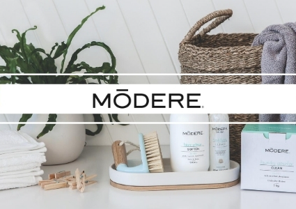 Modere skincare set displayed on a white bathroom counter with a green plant, woven basket, and folded towels.