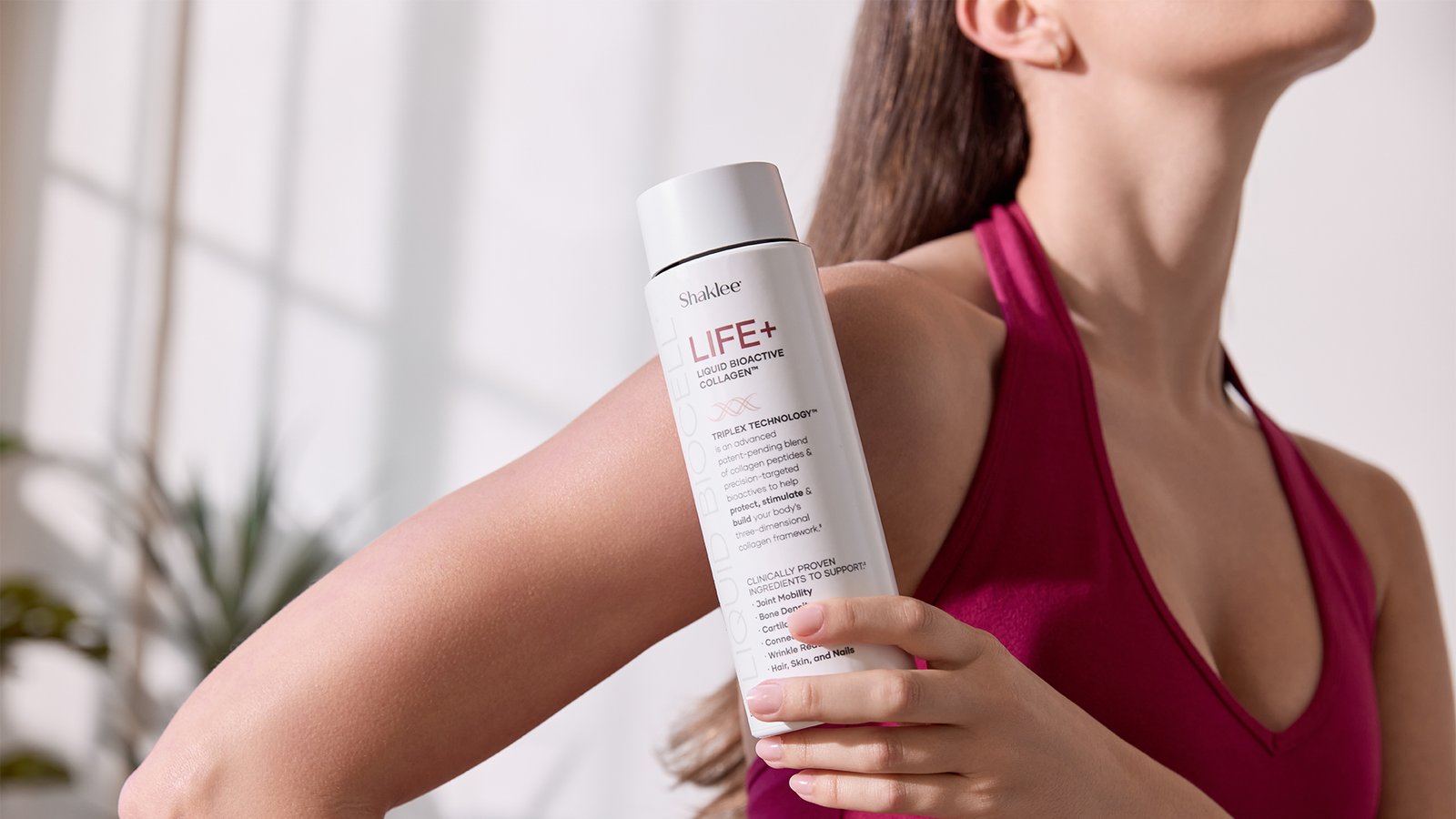 Life+ Model with Bottle in Hand on Shoulder Red Workout Gear - Shop Shaklee Australia Woman in a magenta top holds a white bottle labeled LIFE+ against her shoulder in a bright, airy room.